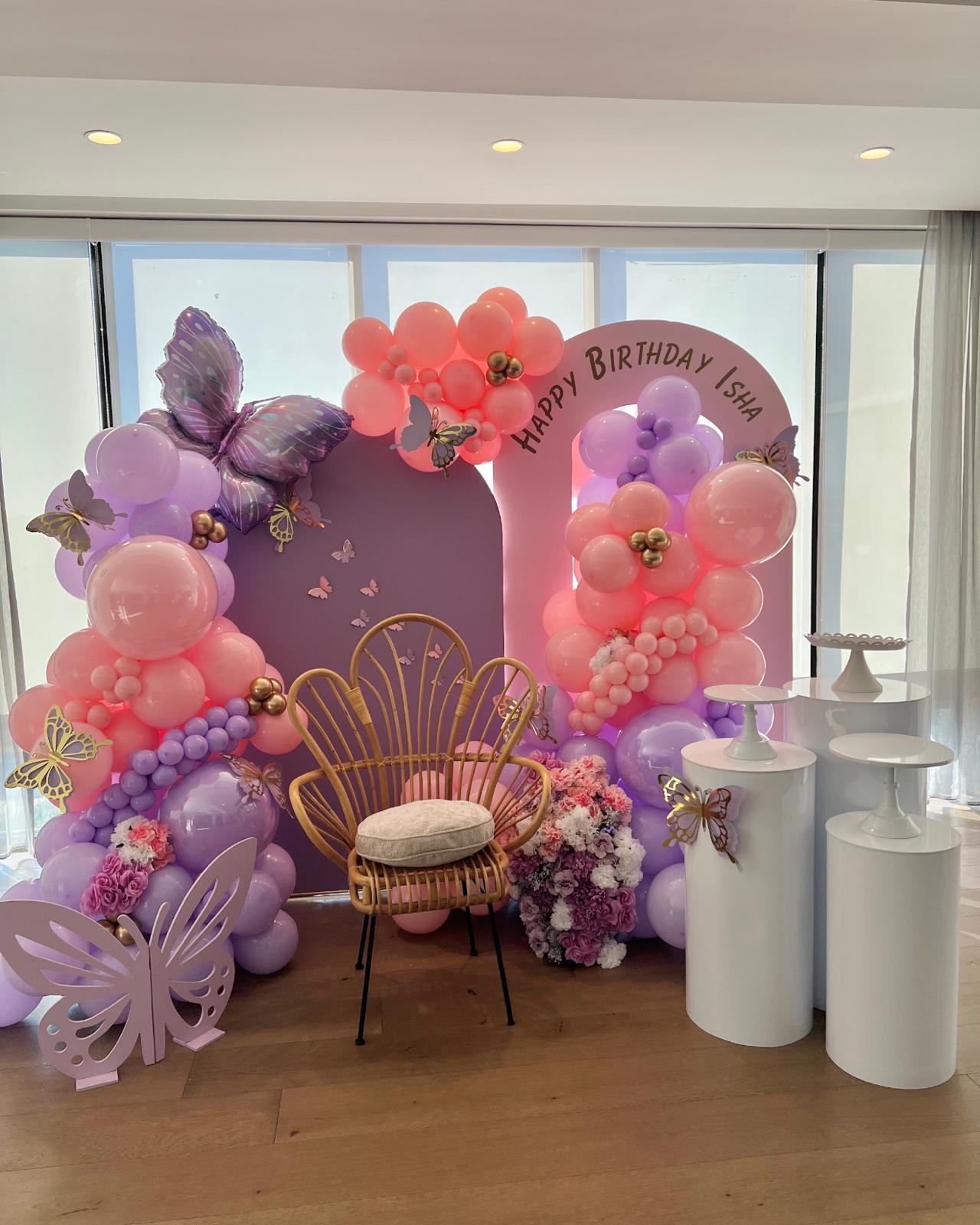 Pink and lilac butterfly themed birthday backdrop with balloon garland and rattan chair